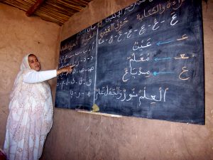 Projekt_Bildung_quer Eine Frau in weißem Gewand steht vor einer Tafel mit arabischen Schriftzeichen. Sie deutet mit einer Hand auf das Geschriebene.