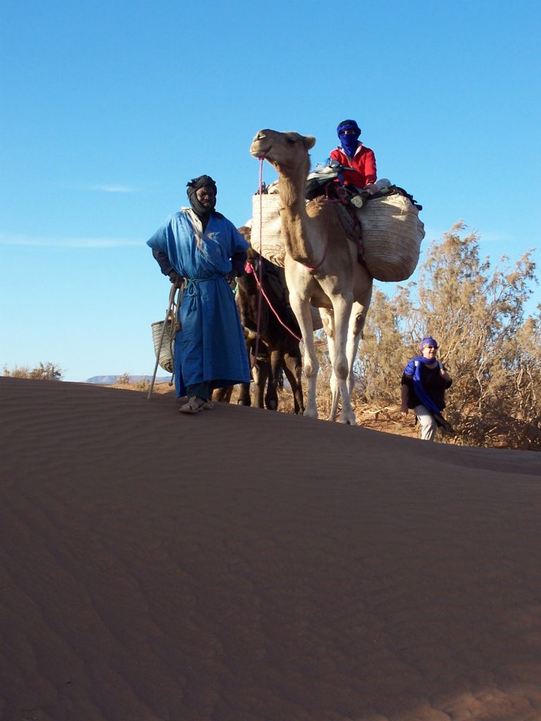 Abdu mit Dromedare und Gäste Ein Tourist reitet auf einem Dromedar. Ein Nomade in blauer Gandura führt das Dromedar an einem Seil über eine Düne.